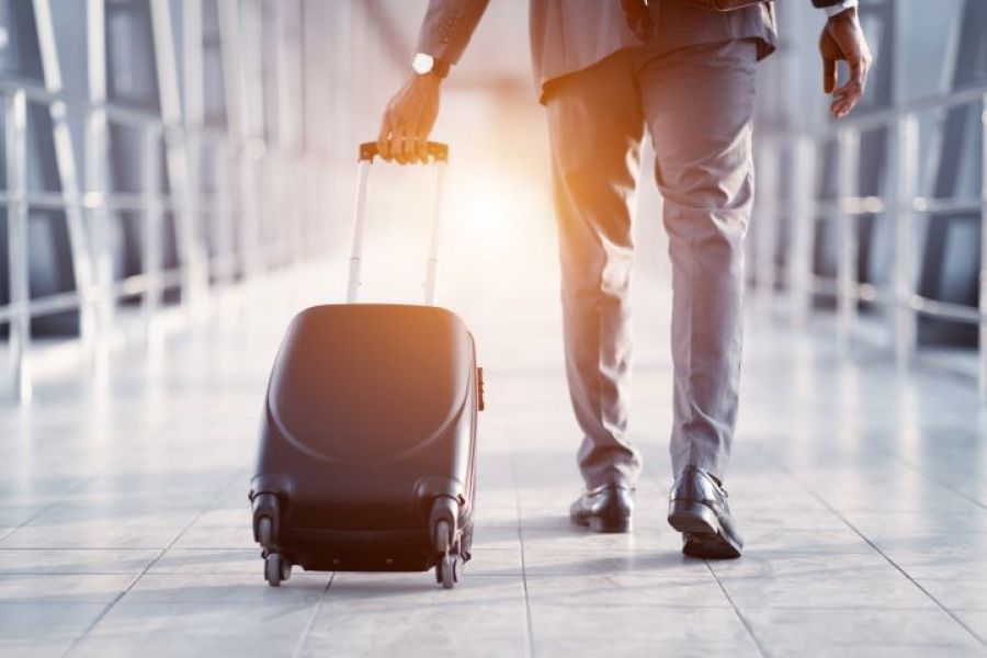 The photo presents a man at the airport, who's going for his trip in front of the rising sun. He holds a black suitcase in his left hand.