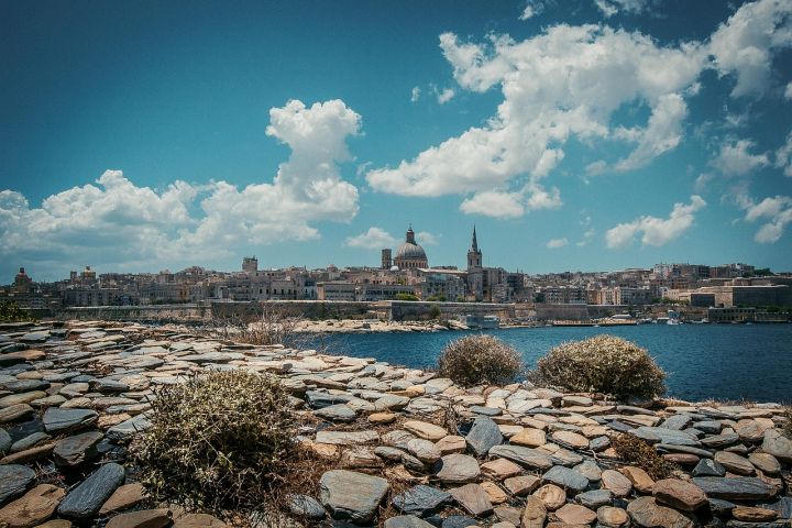 View of the Maltese coastline. In the foreground, the azure waters of the Mediterranean Sea can be seen. In the distance, rocky beaches and old town buildings are visible. 